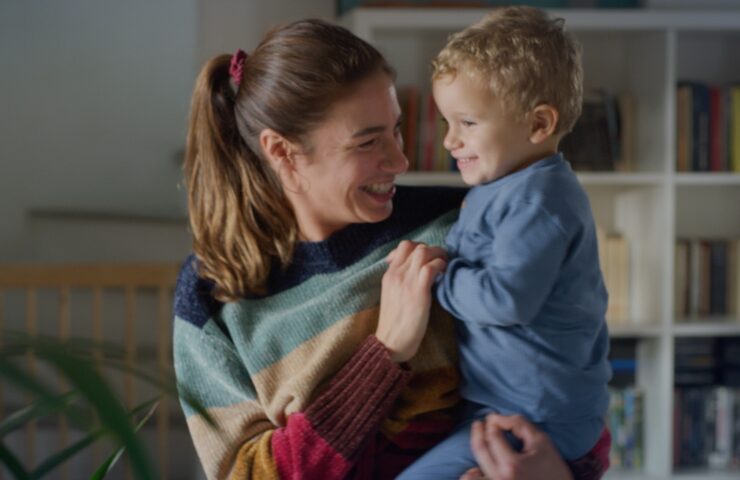 Vrouw met vrolijke glimlach houdt een peuter in haar armen, beiden kijken elkaar liefdevol aan in een huiselijke omgeving.